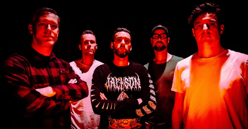A group of five men standing closely together with arms crossed, set against a dark background illuminated by red lighting.