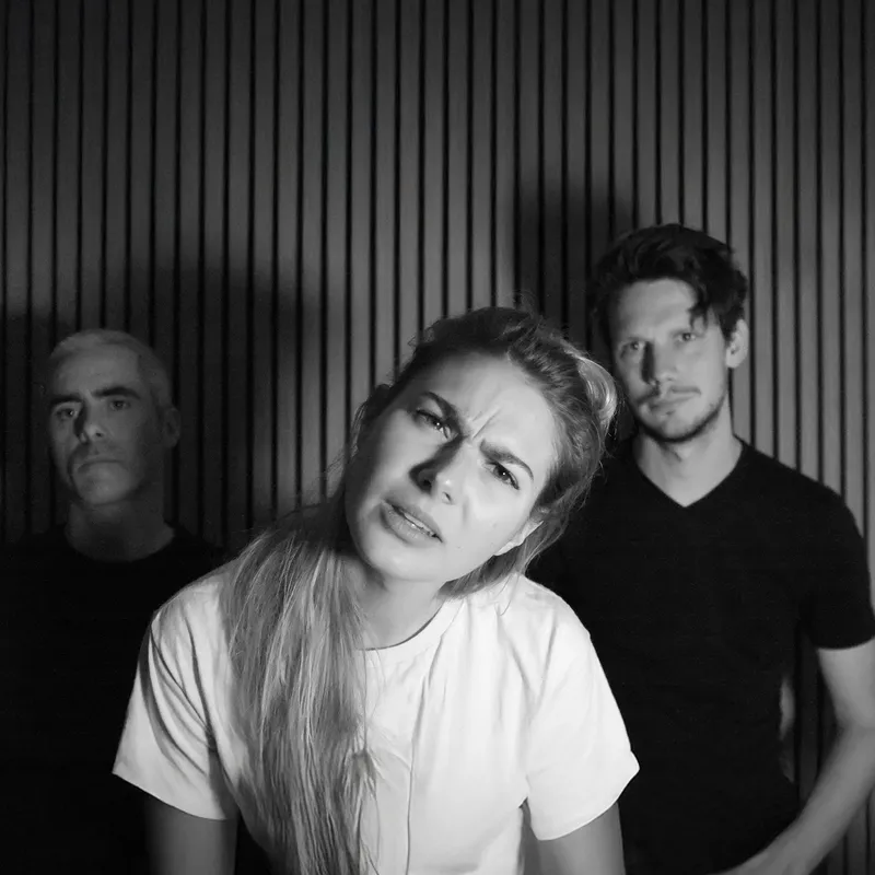 A group of three people posing for a photo in black and white. The woman in front is making a curious expression, while the two men behind her appear serious. The background has vertical lines.