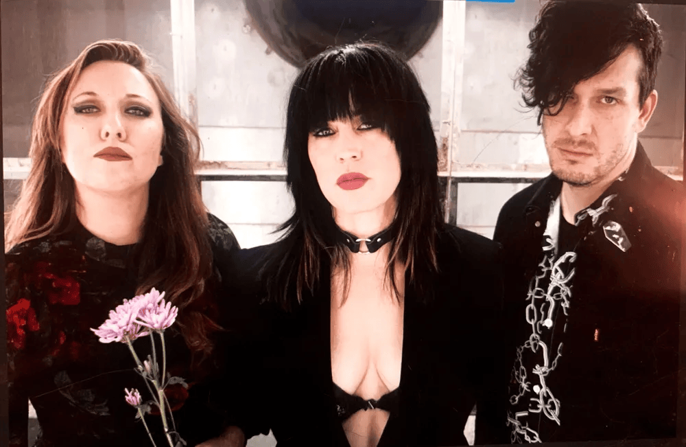 A trio of musicians posing together; two women and one man. One woman holds a flower and wears a floral top, while the other has dark hair and a choker, dressed in black. The man is wearing a patterned shirt. The background is industrial and muted.