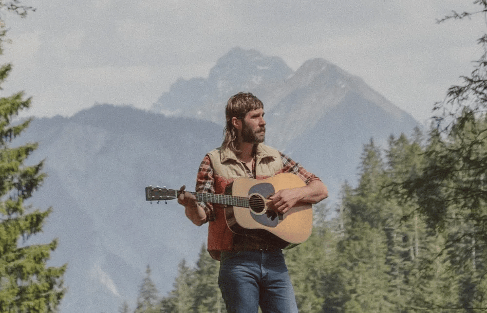 A man with long hair and a beard stands in a mountainous outdoor setting, playing an acoustic guitar. He is wearing a plaid shirt and a vest, surrounded by trees and a scenic backdrop of mountains.