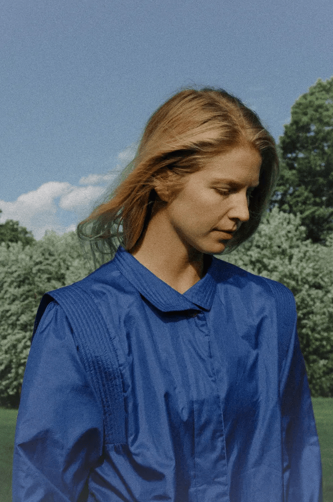 A woman in a blue jacket stands outdoors, looking down. She has long hair and is surrounded by greenery and a clear blue sky.