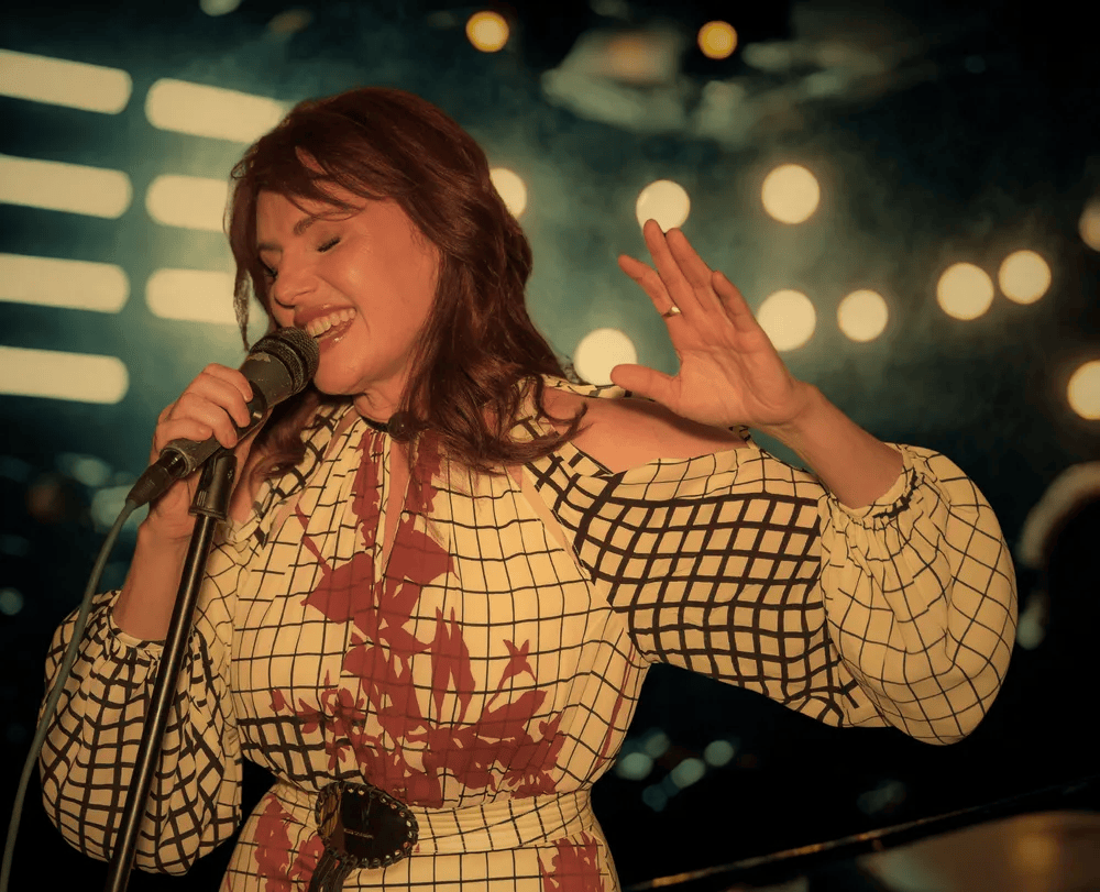 A singer performing on stage, wearing a patterned dress and holding a microphone, with a joyful expression and stage lights in the background.