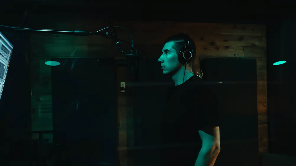 A person wearing headphones stands in a recording studio, facing a microphone, with a computer screen displaying audio editing software in the background.
