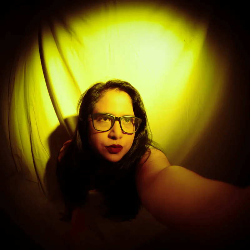 A woman with long black hair and glasses poses under a warm yellow light backdrop, looking thoughtfully to the side.