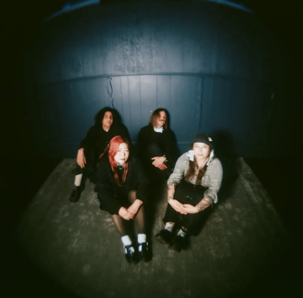 Four individuals sitting on a wooden floor in a dimly lit space, with a dark blue wall in the background. They have a relaxed posture, with two wearing dark clothing and one with red hair. The atmosphere appears casual and intimate.