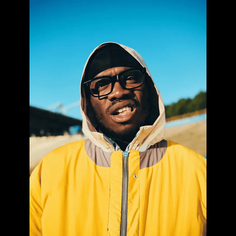 A man wearing a yellow jacket and glasses stands outdoors with a clear blue sky in the background, looking directly at the camera.
