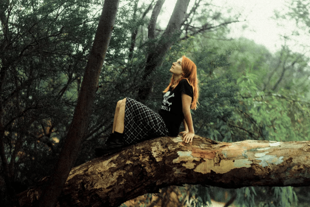 A young woman with red hair sits on a tree branch in a lush green forest, wearing a black t-shirt and a plaid skirt, gazing upward.