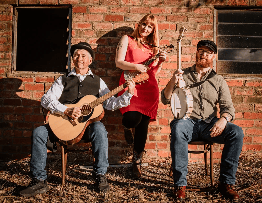 A trio of musicians posing outdoors in front of a brick wall. One male musician plays an acoustic guitar, while a female musician holds a ukulele, and another male musician sits with a banjo. They are dressed casually, with a mix of formal and informal attire.