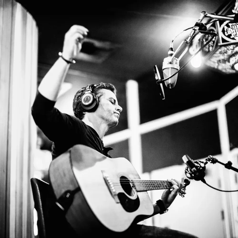 A musician wearing headphones plays an acoustic guitar in a recording studio, with a microphone positioned above him.