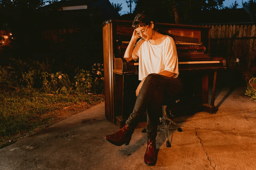 A woman sitting next to a wooden piano in a softly lit outdoor setting, wearing a relaxed outfit and stylish boots.