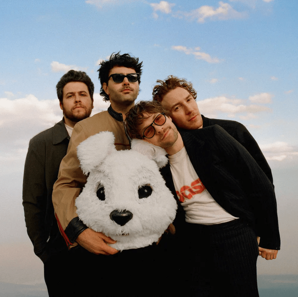 A group of four men posing outdoors, with a light blue sky in the background. One of the men is holding a large, fluffy white bear head. The others are standing close together, showcasing a casual and relaxed vibe.