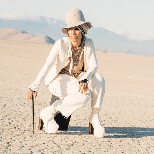 A woman in high-heeled boots and a stylish outfit sits on a sandy landscape, holding a cane, with mountains in the background.