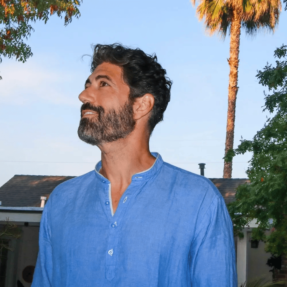 A man with a beard in a blue shirt looking up towards the sky, with palm trees and greenery in the background.