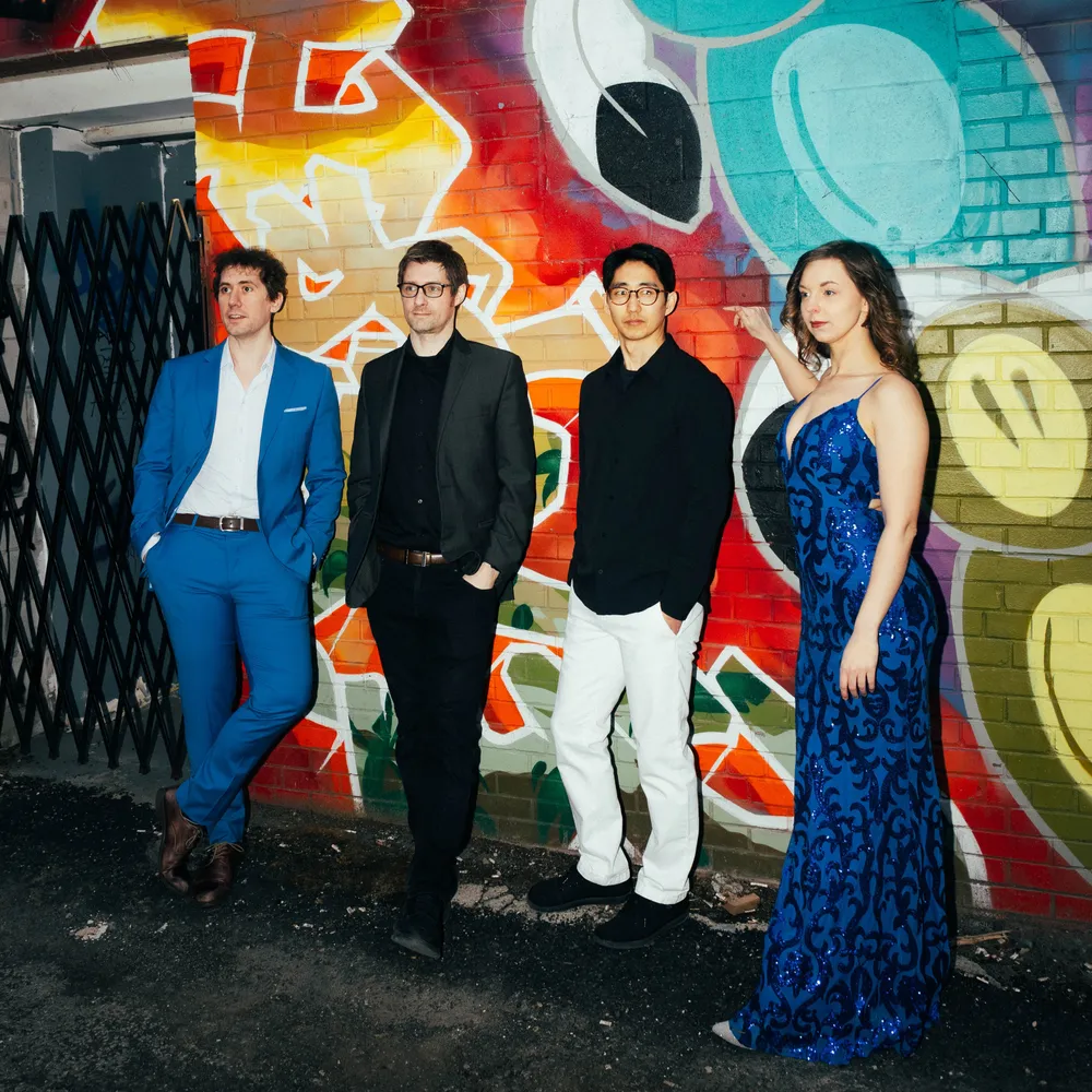 Four fashionable individuals posing against a colorful graffiti wall. The first person wears a blue suit, the second wears a black suit, the third wears a black shirt, and the fourth wears a blue sequined dress.